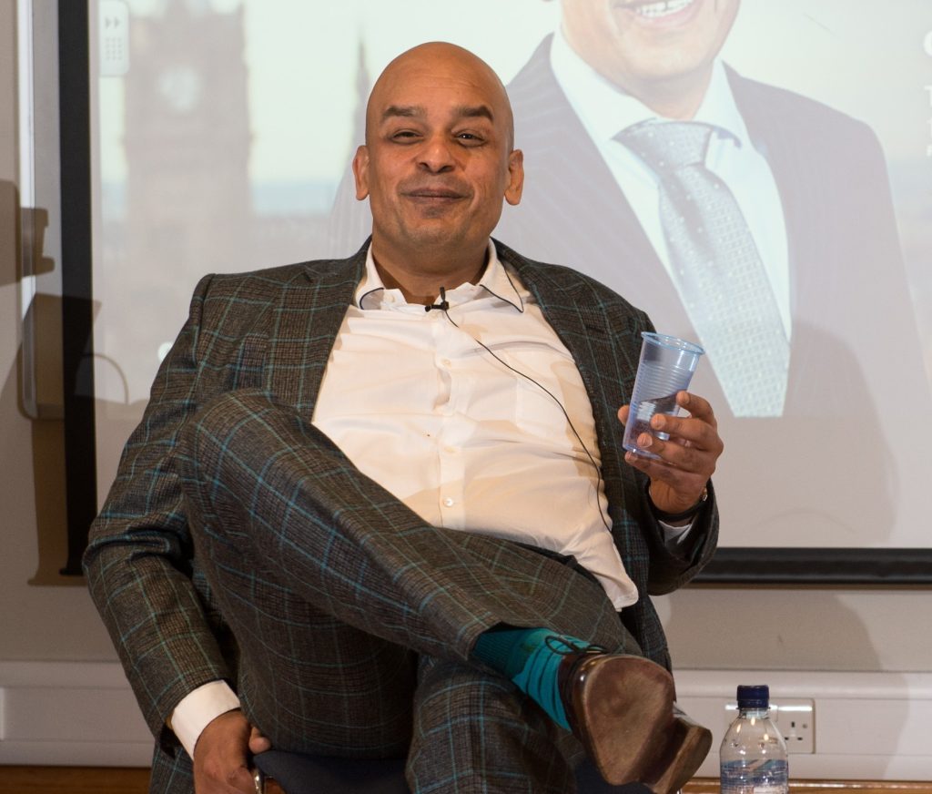 Shaf Rasul seated in a suit with legs crossed, holding a glass of water, illustrating a webinar discussion