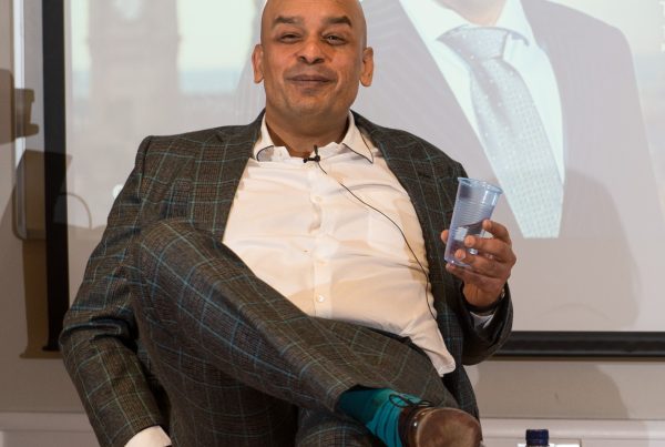 Shaf Rasul seated in a suit with legs crossed, holding a glass of water, illustrating a webinar discussion