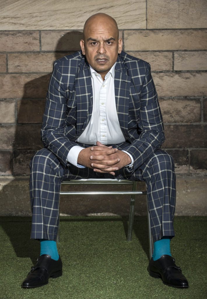 Shaf Rasul sitting on a chair in a checked suit with blue socks, against a stone wall backdrop.