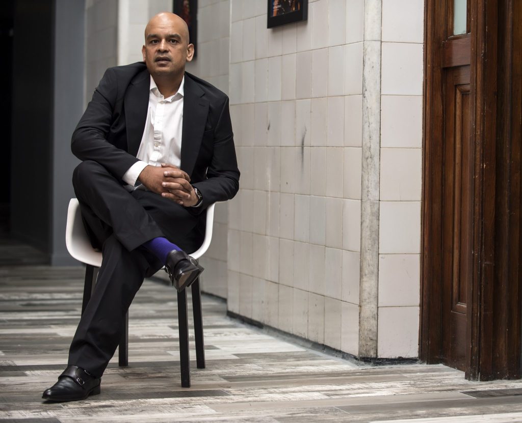 Shaf Rasul sitting cross-legged on a chair in a modern interior with tiled walls and wooden doors.