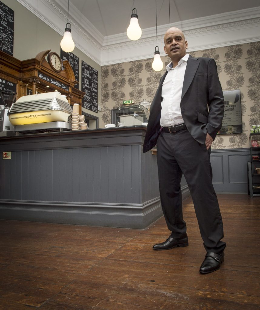 Shaf Rasul standing confidently inside a cafe in Edinburgh, wearing a dark suit and white shirt.