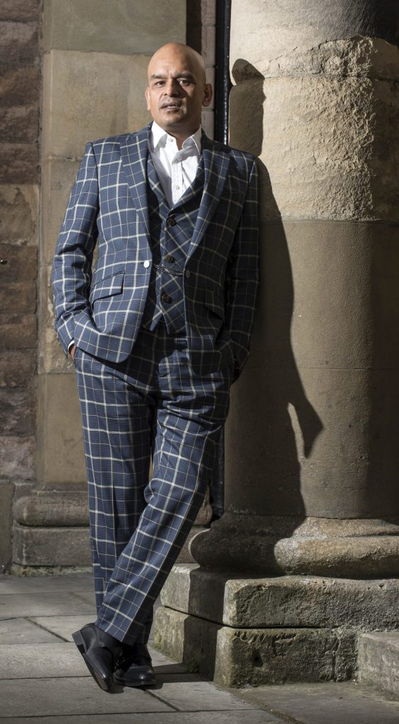 Shaf Rasul standing against large stone columns in Edinburgh wearing a blue checked three-piece suit.