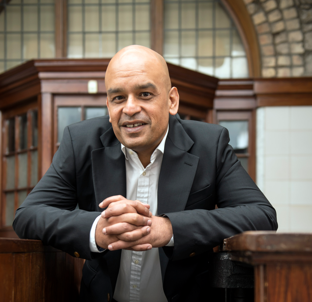 Shaf Rasul leaning on a wooden surface, smiling in a business portrait with a wood and glass backdrop.