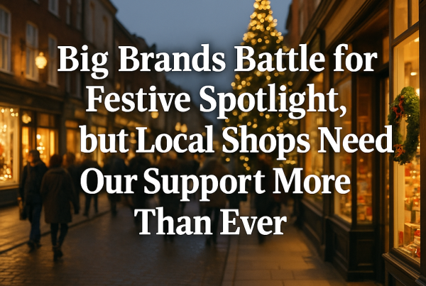 A square-format photo of a warmly lit high-street at dusk during the Christmas season: shop windows glow with festive decorations, a decorated tree stands in the distance, and crowds stroll past. Superimposed in large white serif letters is the headline: “Big Brands Battle for Festive Spotlight, but Local Shops Need Our Support More Than Ever.”